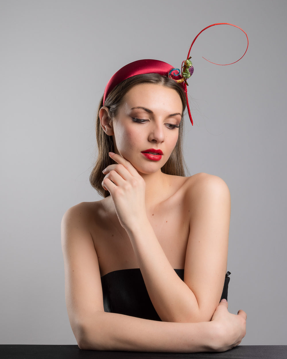 The female model is posing, wearing
Bukelamun,  the red satin padded headband featuring a colorful jeweled chameleon embellishment with a curled tail and a thin red sculptural wire detail extending upward.