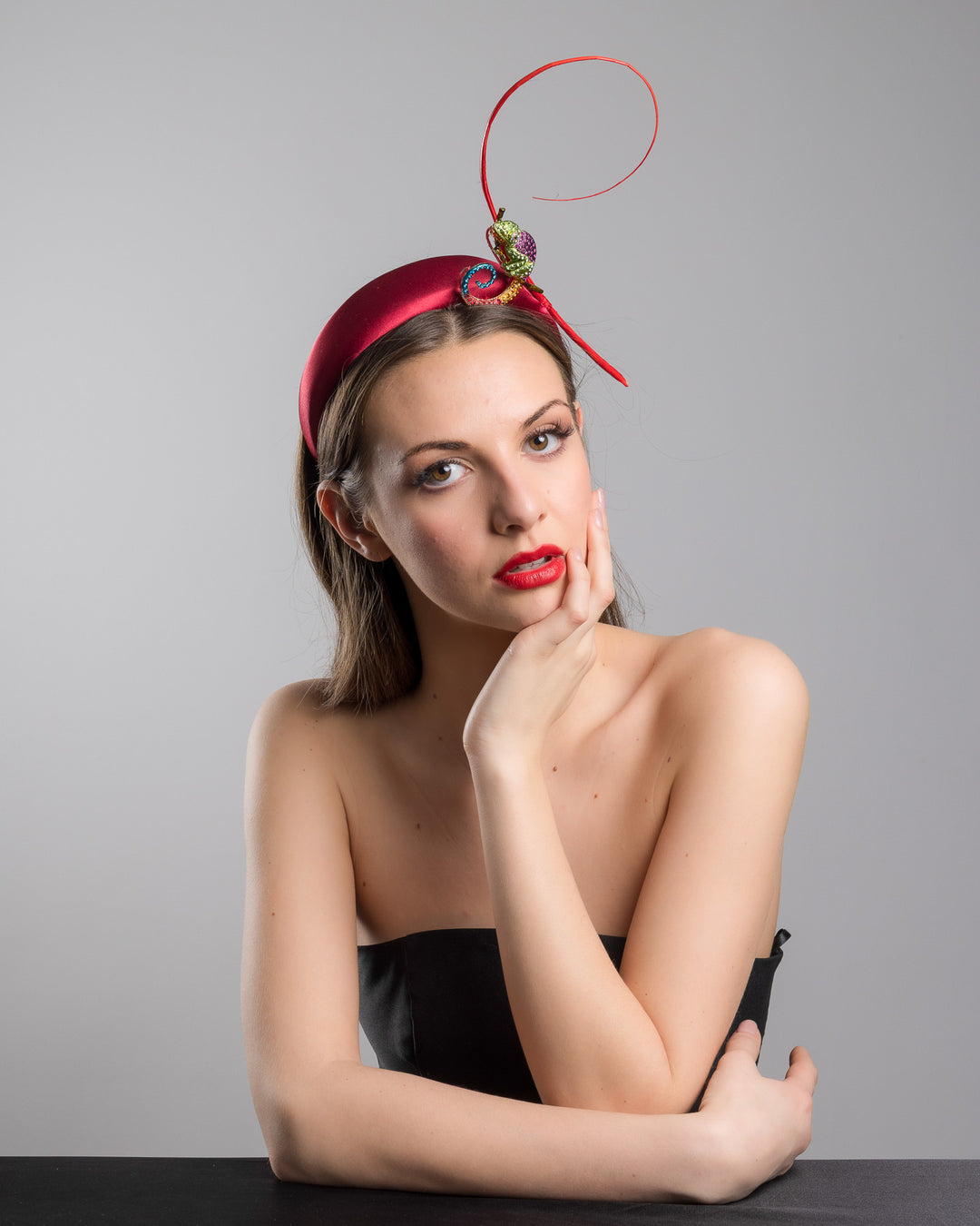 The female model is posing, wearing
Bukelamun,  the red satin padded headband featuring a colorful jeweled chameleon embellishment with a curled tail and a thin red sculptural wire detail extending upward.