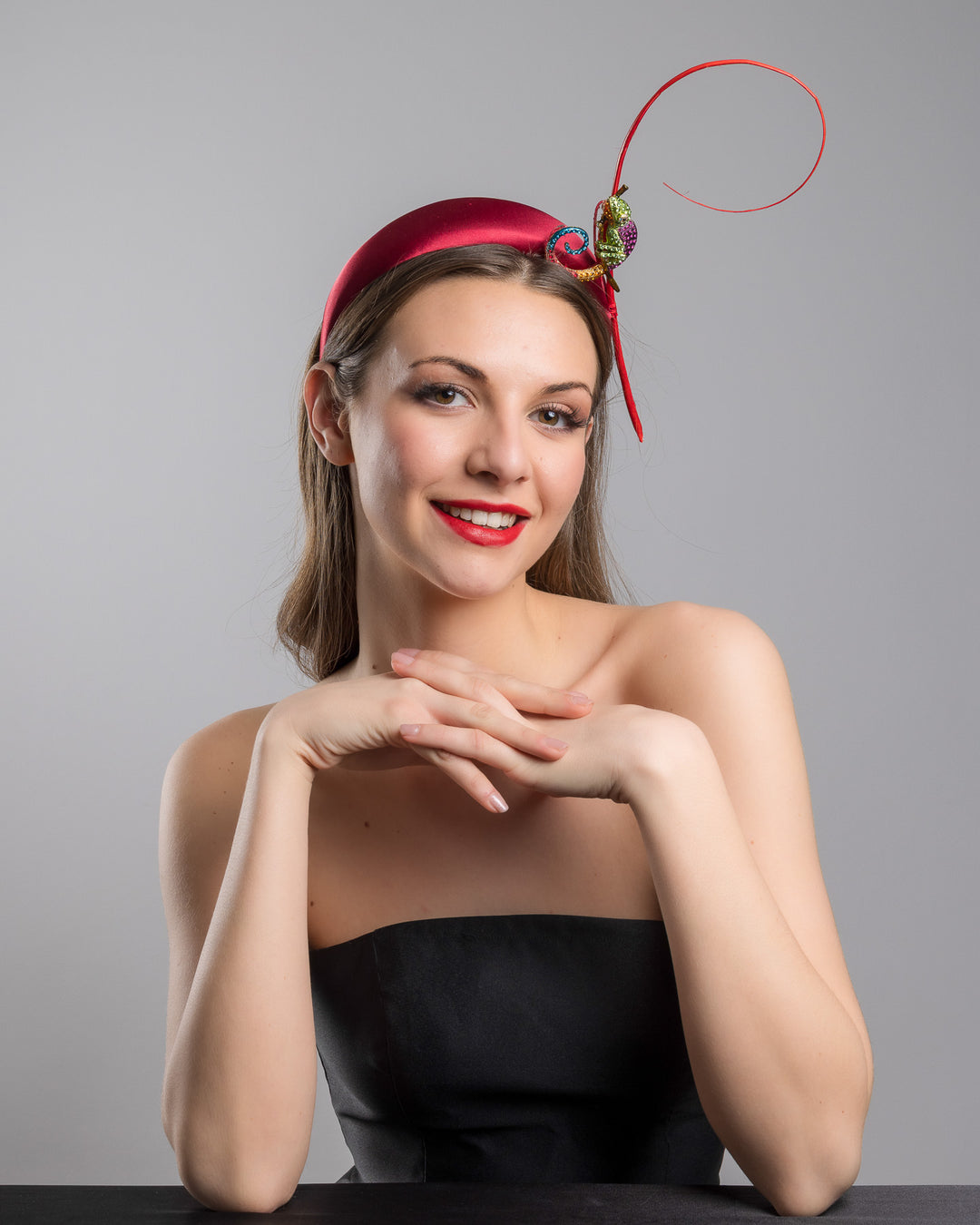 The female model is posing, wearing
Bukelamun,  the red satin padded headband featuring a colorful jeweled chameleon embellishment with a curled tail and a thin red sculptural wire detail extending upward.