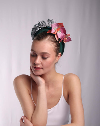 The model is wearing a deep green velvet high headband. The top of the headband is adorned with a peacock feather placed like an arch. On the left side end of the feather, two orchid flowers in flowing light purple, orange, and pink coloured. 