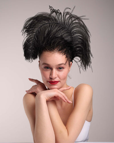 The model is wearing a large black ostrich feather adorned with black crystals in the centre. Half of the feathers fall on top of her head, while the part above the crystals that are lined up flies in the air. The arced black ostrich feather is fitted to the head on a hidden headband. This piece is perfect for high-fashion events as well as costume parties.