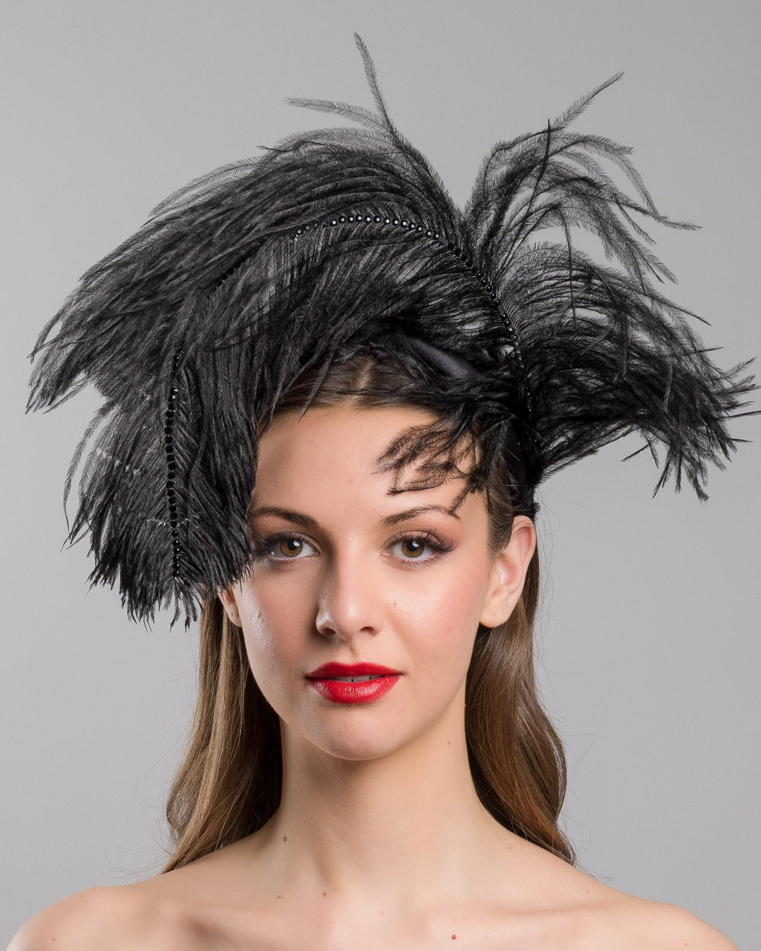 Model is showcasing the front side close up af a luxury black sateen headband fitted with a large black ostrich feather on one side curving on top of the headband. The ostrich feather is adorned with black crystals in the centre. The ostrich feather on the headband creates an illusion of standing in the air. Made by Merve Bayindir Millinery.
