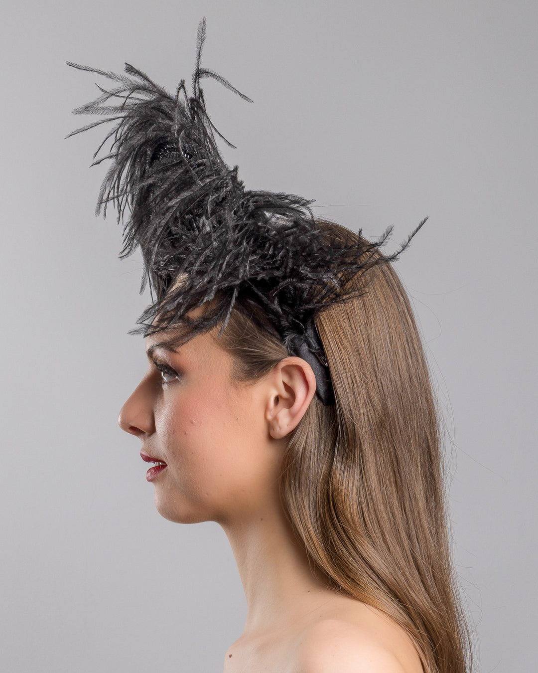 Model is showcasing the left side close up af a luxury black sateen headband fitted with a large black ostrich feather on one side curving on top of the headband. The ostrich feather is adorned with black crystals in the centre. The ostrich feather on the headband creates an illusion of standing in the air. Made by Merve Bayindir Millinery.