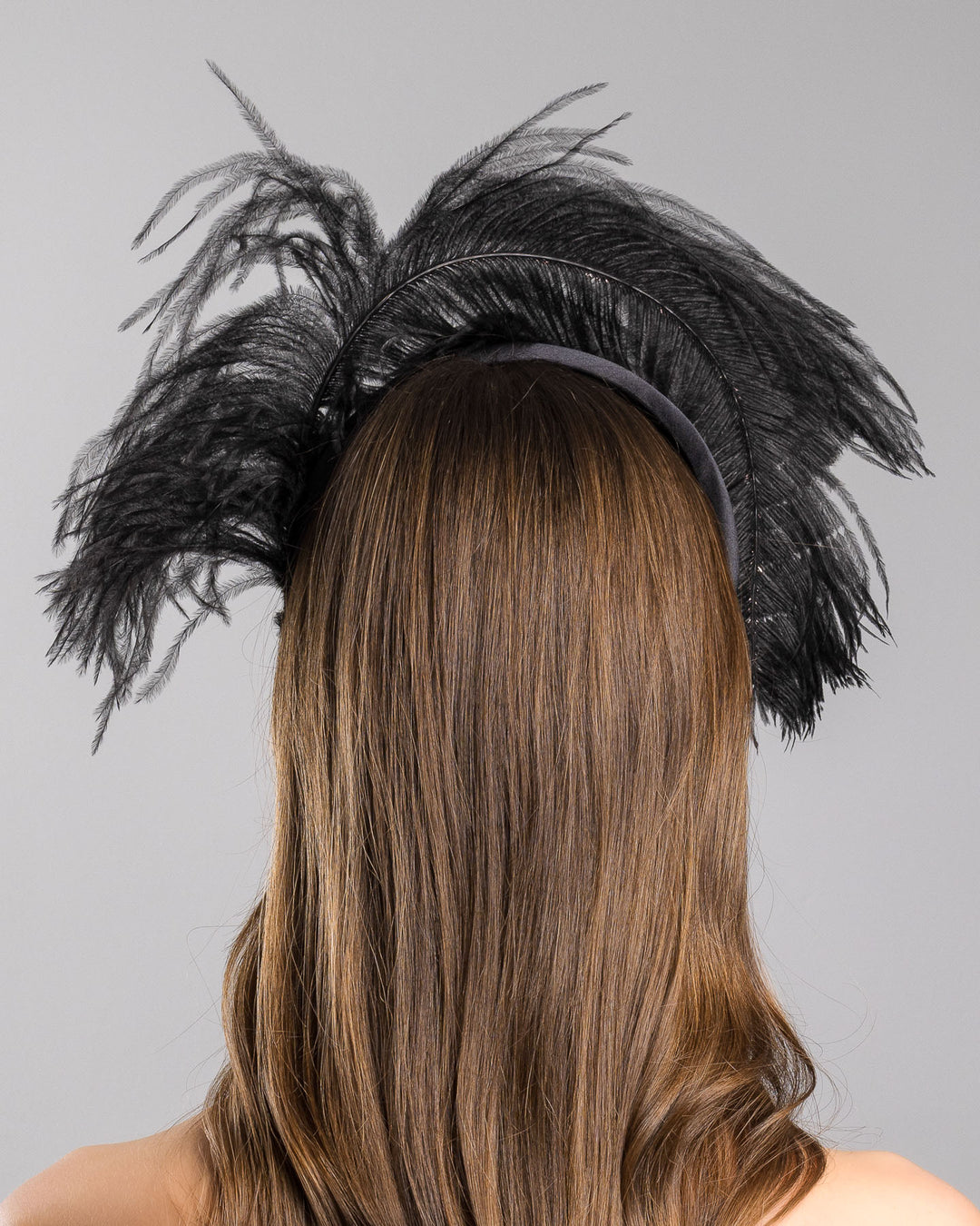 Model is showcasing the back side close up af a luxury black sateen headband fitted with a large black ostrich feather on one side curving on top of the headband. The ostrich feather is adorned with black crystals in the centre. The ostrich feather on the headband creates an illusion of standing in the air. Made by Merve Bayindir Millinery.