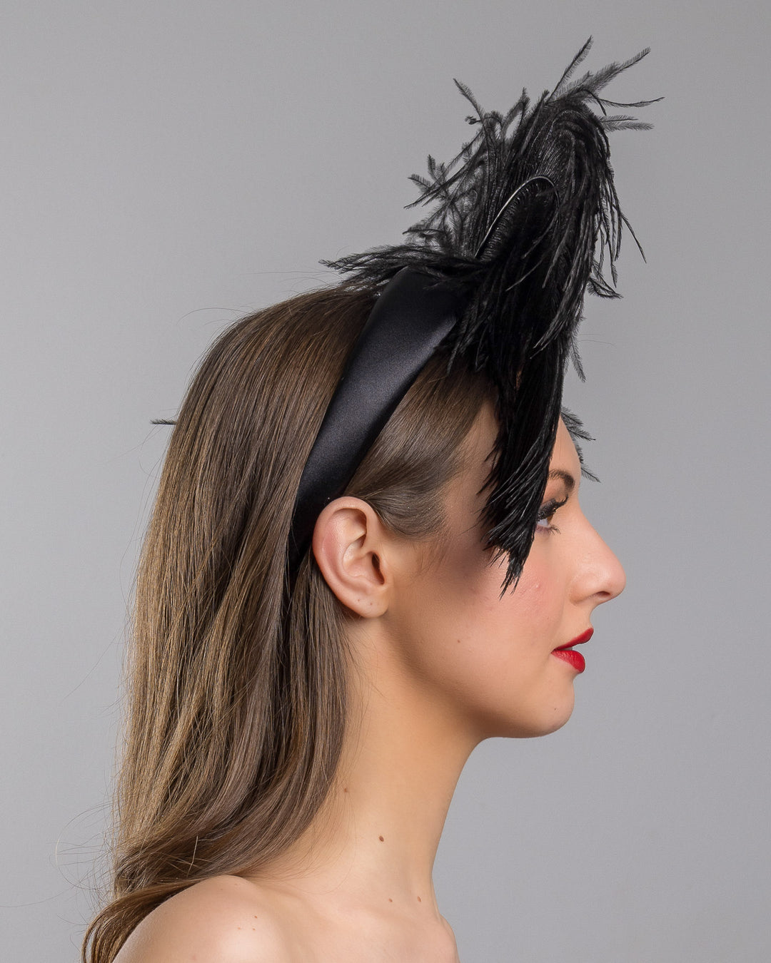 Model is showcasing the right side close up af a luxury black sateen headband fitted with a large black ostrich feather on one side curving on top of the headband. The ostrich feather is adorned with black crystals in the centre. The ostrich feather on the headband creates an illusion of standing in the air. Made by Merve Bayindir Millinery.