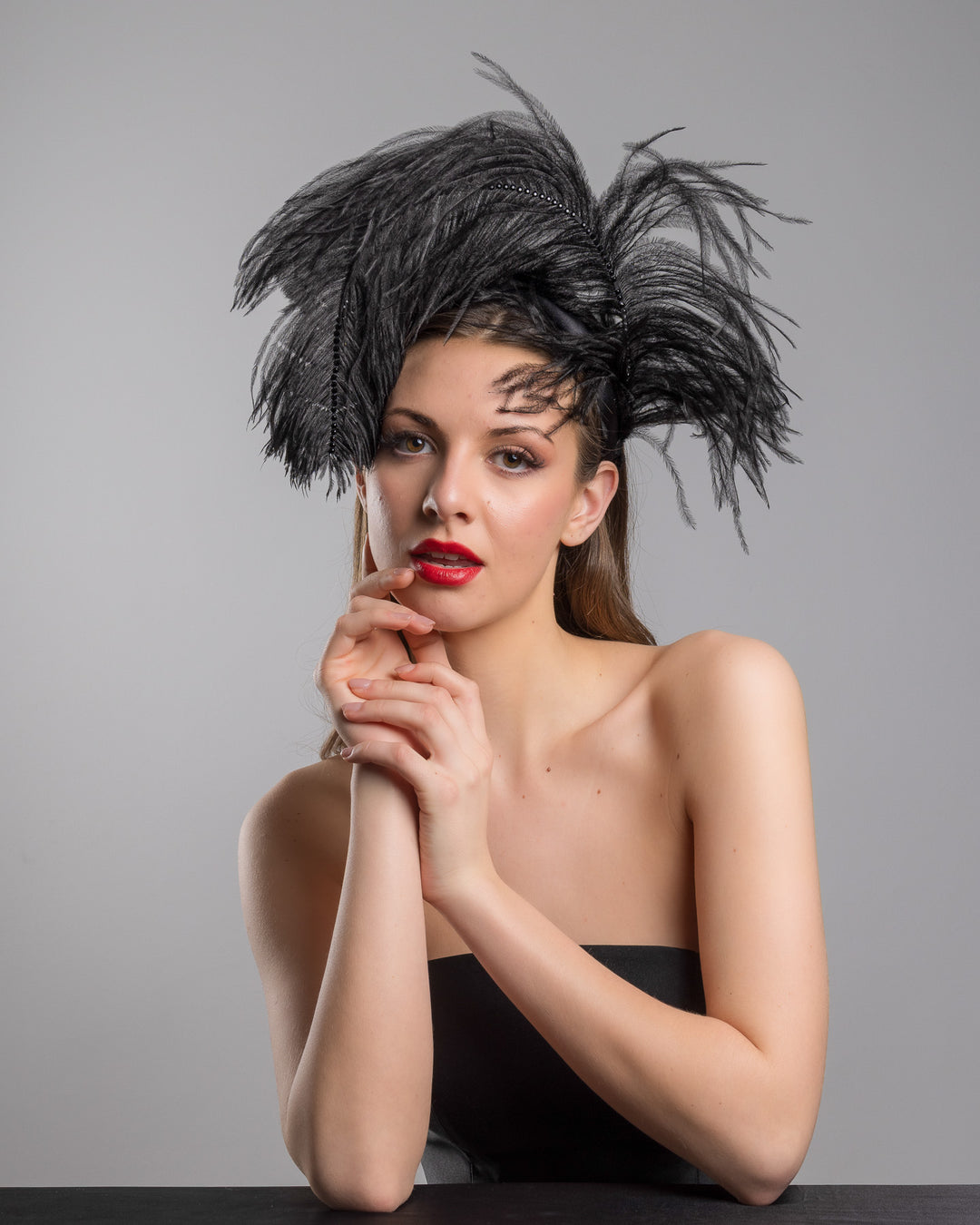 Female model is posing wearing Tango, a luxury black sateen headband fitted with a large black ostrich feather on one side curving on top of the headband. The ostrich feather is adorned with black crystals in the centre. The ostrich feather on the headband creates an illusion of standing in the air. Made by Merve Bayindir Millinery.