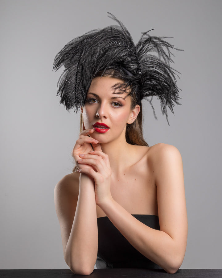 Female model is posing wearing Tango, a luxury black sateen headband fitted with a large black ostrich feather on one side curving on top of the headband. The ostrich feather is adorned with black crystals in the centre. The ostrich feather on the headband creates an illusion of standing in the air. Made by Merve Bayindir Millinery.