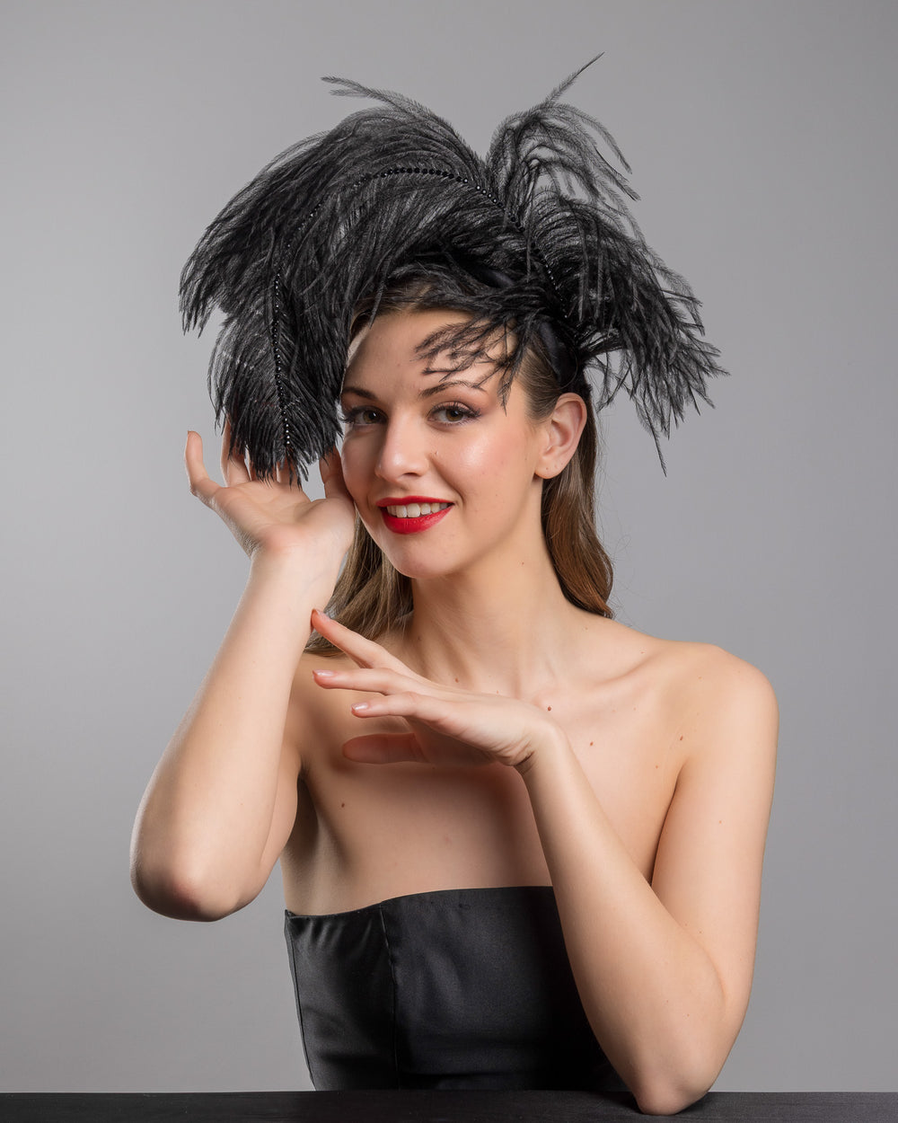 Female model is posing wearing Tango, a luxury black sateen headband fitted with a large black ostrich feather on one side curving on top of the headband. The ostrich feather is adorned with black crystals in the centre. The ostrich feather on the headband creates an illusion of standing in the air. Made by Merve Bayindir Millinery.