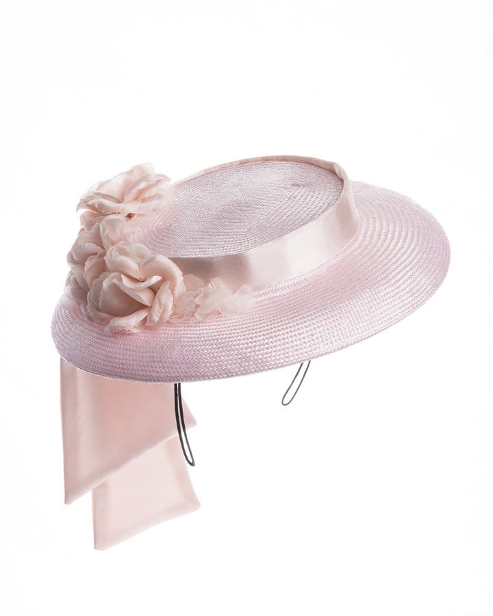 Blush pink mini boater hat crafted from woven straw, trimmed with a satin band and adorned with soft pink floral accents and a flowing satin ribbon at the back, mounted on a slim headband. Designed and made by Merve Bayindir Millinery.
