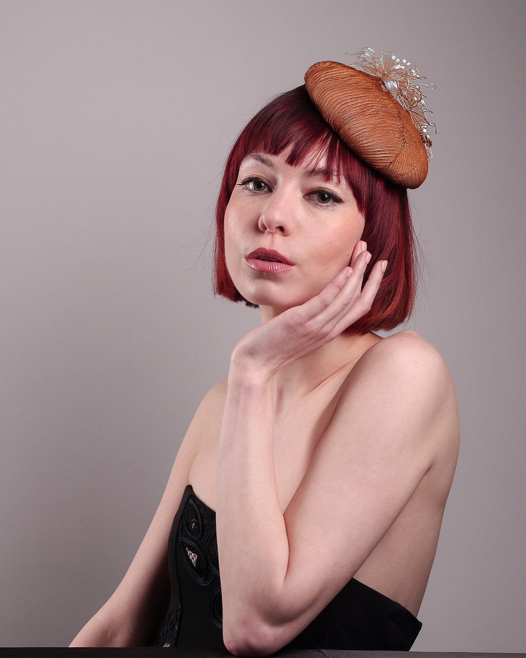 The model is showcasing the front of the Belle Fascinator button-shaped hand-formed pillbox hat in a warm cinnamon tone, wrapped in finely pleated organza for rich texture and depth. Sitting neatly on the crown, it’s finished with delicate beads and translucent resin petals. Designed by Merve Bayindir. 