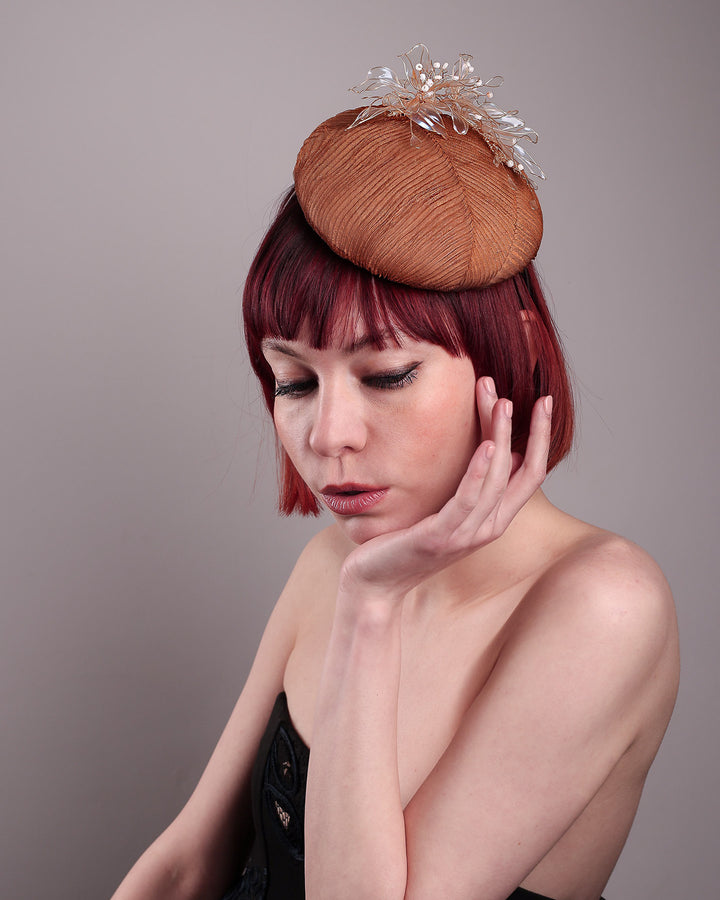 The model is showcasing the close up top of the  Belle Fascinator button-shaped hand-formed pillbox hat in a warm cinnamon tone, wrapped in finely pleated organza for rich texture and depth. Sitting neatly on the crown, it’s finished with delicate beads and translucent resin petals. Designed by Merve Bayindir. 