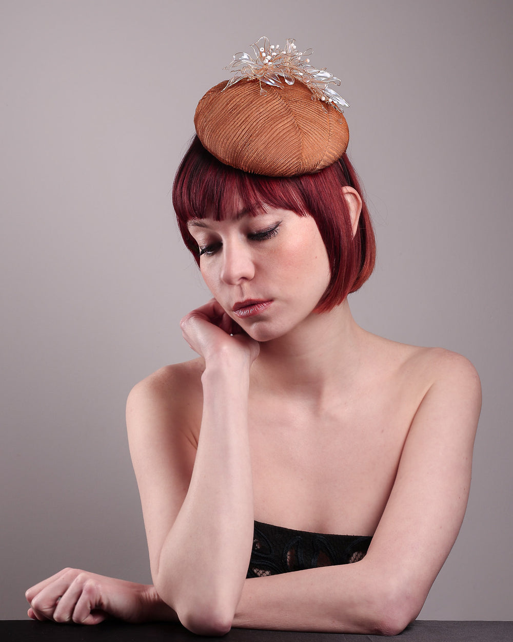 The model is showcasing the above angle of the Belle Fascinator button-shaped hand-formed pillbox hat in a warm cinnamon tone, wrapped in finely pleated organza for rich texture and depth. Sitting neatly on the crown, it’s finished with delicate beads and translucent resin petals. Designed by Merve Bayindir. 