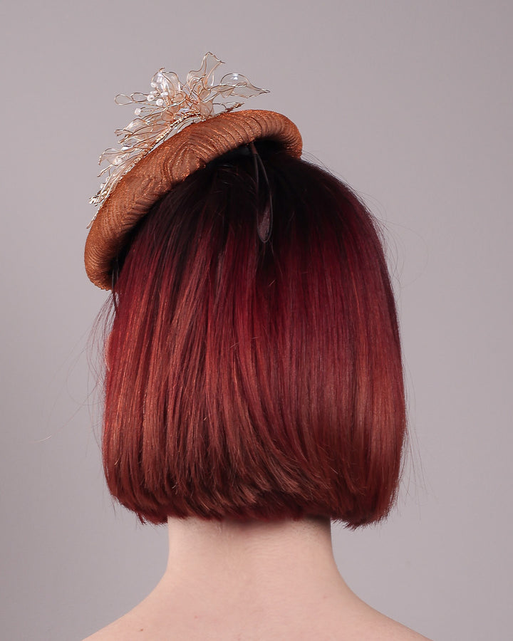 The model is showcasing the back of the Belle Fascinator button-shaped hand-formed pillbox hat in a warm cinnamon tone, wrapped in finely pleated organza for rich texture and depth. Sitting neatly on the crown, it’s finished with delicate beads and translucent resin petals. Designed by Merve Bayindir. 