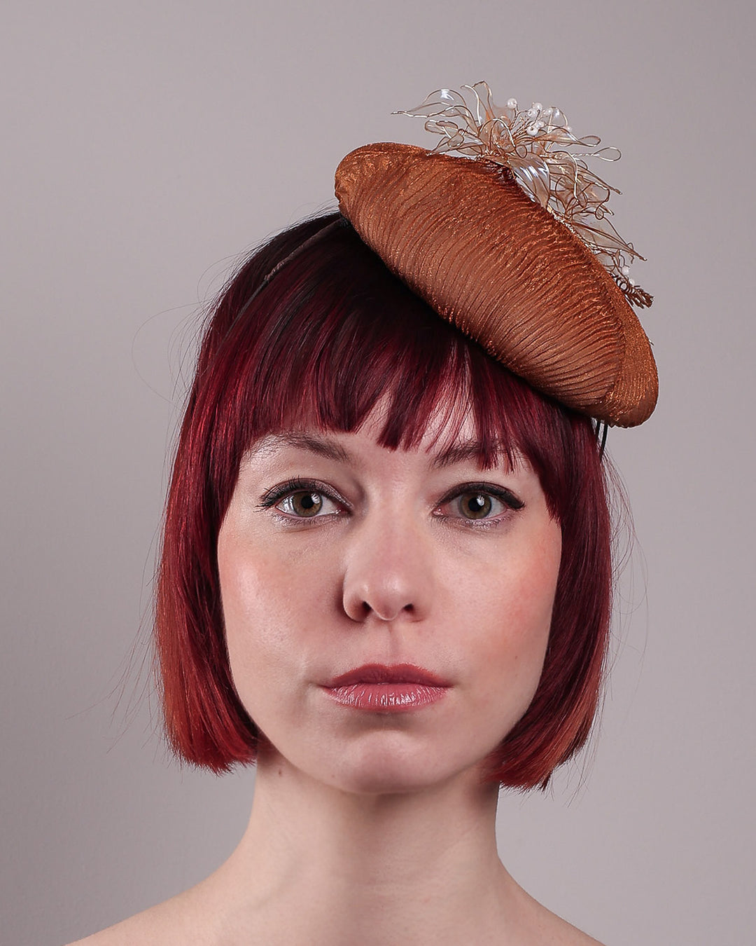 The model is showcasing the front close up of the Belle Fascinator button-shaped hand-formed pillbox hat in a warm cinnamon tone, wrapped in finely pleated organza for rich texture and depth. Sitting neatly on the crown, it’s finished with delicate beads and translucent resin petals. Designed by Merve Bayindir. 