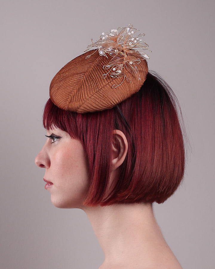 The model is showcasing theleft side close up of the Belle Fascinator button-shaped hand-formed pillbox hat in a warm cinnamon tone, wrapped in finely pleated organza for rich texture and depth. Sitting neatly on the crown, it’s finished with delicate beads and translucent resin petals. Designed by Merve Bayindir. 