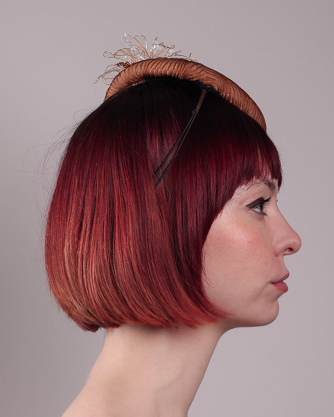 The model is showcasing the right side close up of the Belle Fascinator button-shaped hand-formed pillbox hat in a warm cinnamon tone, wrapped in finely pleated organza for rich texture and depth. Sitting neatly on the crown, it’s finished with delicate beads and translucent resin petals. Designed by Merve Bayindir. 