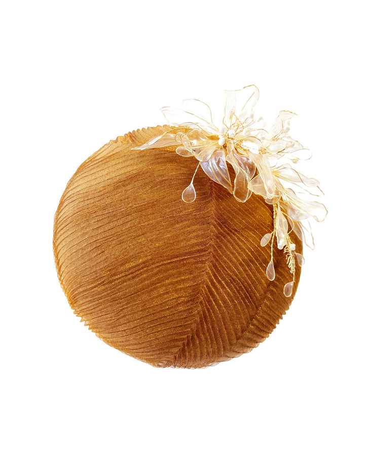 The still life picture of the Belle Fascinator button-shaped hand-formed pillbox hat in a warm cinnamon tone, wrapped in finely pleated organza for rich texture and depth. Sitting neatly on the crown, it’s finished with delicate beads and translucent resin petals. Designed by Merve Bayindir. 