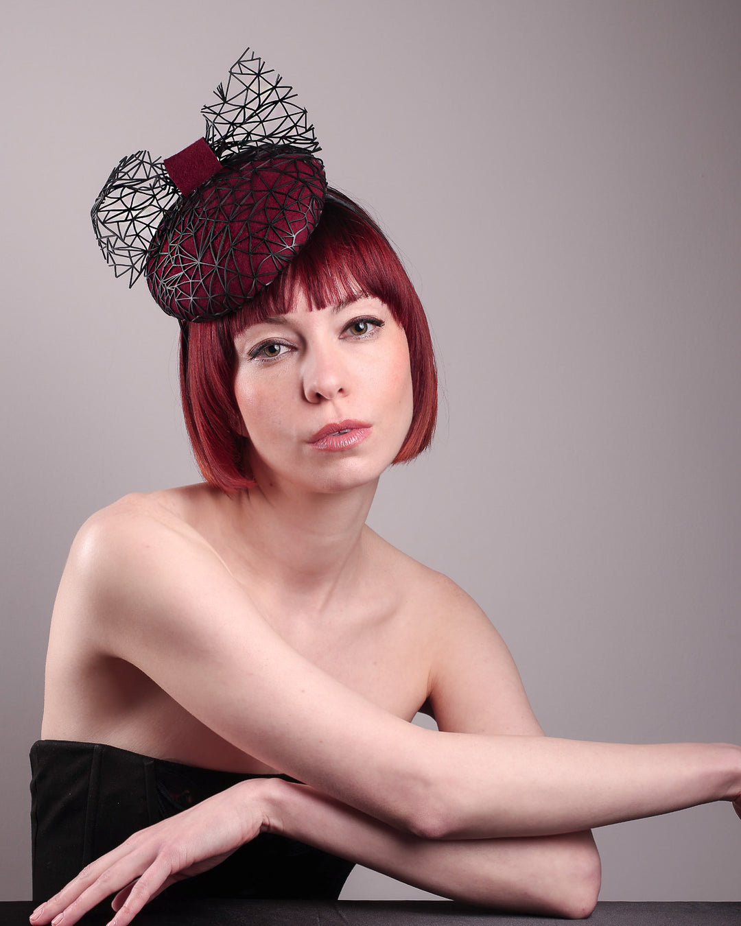 The model showcases the front visual of Bijou, featuring a handcrafted raffia fascinator in rich burgundy felt, with button shape. Its structured base is overlaid with an angular black netting design and crowned with an architectural bow that fuses avant-garde with elegance. Designed by Merve Bayindir.