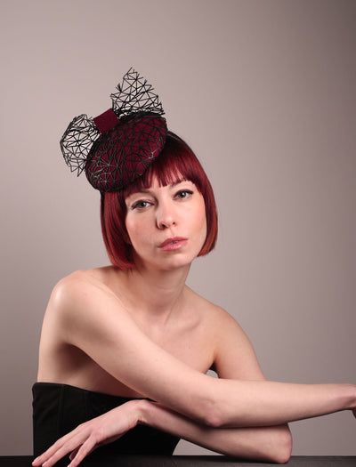 The model showcases the front visual of Bijou, featuring a handcrafted raffia fascinator in rich burgundy felt, with button shape. Its structured base is overlaid with an angular black netting design and crowned with an architectural bow that fuses avant-garde with elegance. Designed by Merve Bayindir.