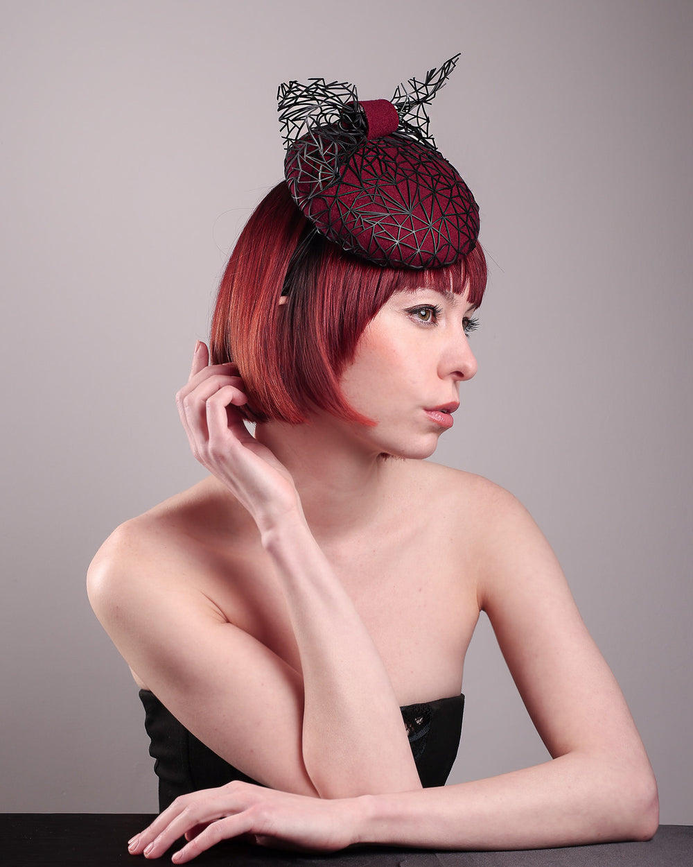 The model showcases the right front angle of Bijou, featuring a handcrafted raffia fascinator in rich burgundy felt, with button shape. Its structured base is overlaid with an angular black netting design and crowned with an architectural bow that fuses avant-garde with elegance. Designed by Merve Bayindir.