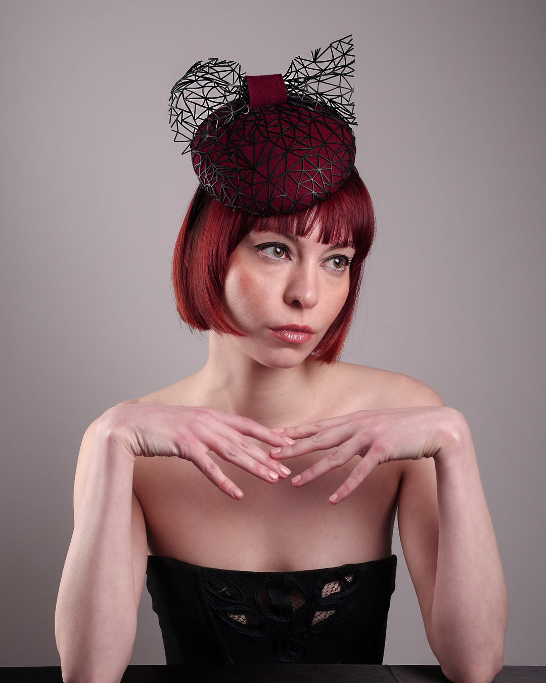 The model showcases the front above angle of Bijou, featuring a handcrafted raffia fascinator in rich burgundy felt, with button shape. Its structured base is overlaid with an angular black netting design and crowned with an architectural bow that fuses avant-garde with elegance. Designed by Merve Bayindir.