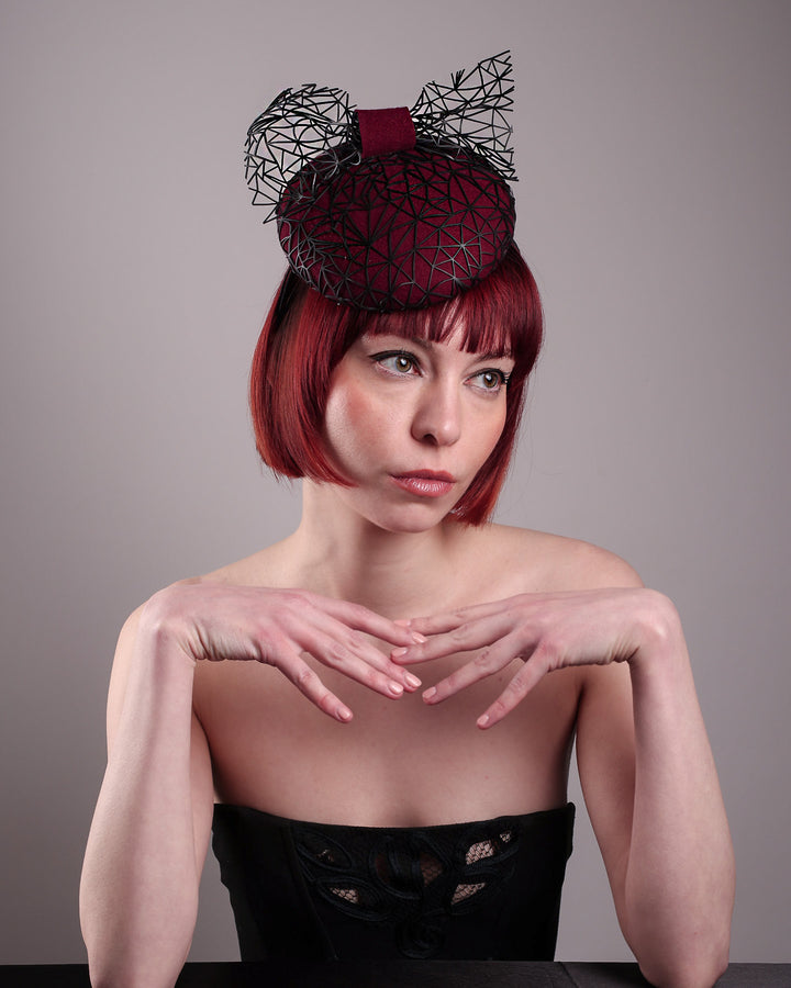 The model showcases the front above angle of Bijou, featuring a handcrafted raffia fascinator in rich burgundy felt, with button shape. Its structured base is overlaid with an angular black netting design and crowned with an architectural bow that fuses avant-garde with elegance. Designed by Merve Bayindir.