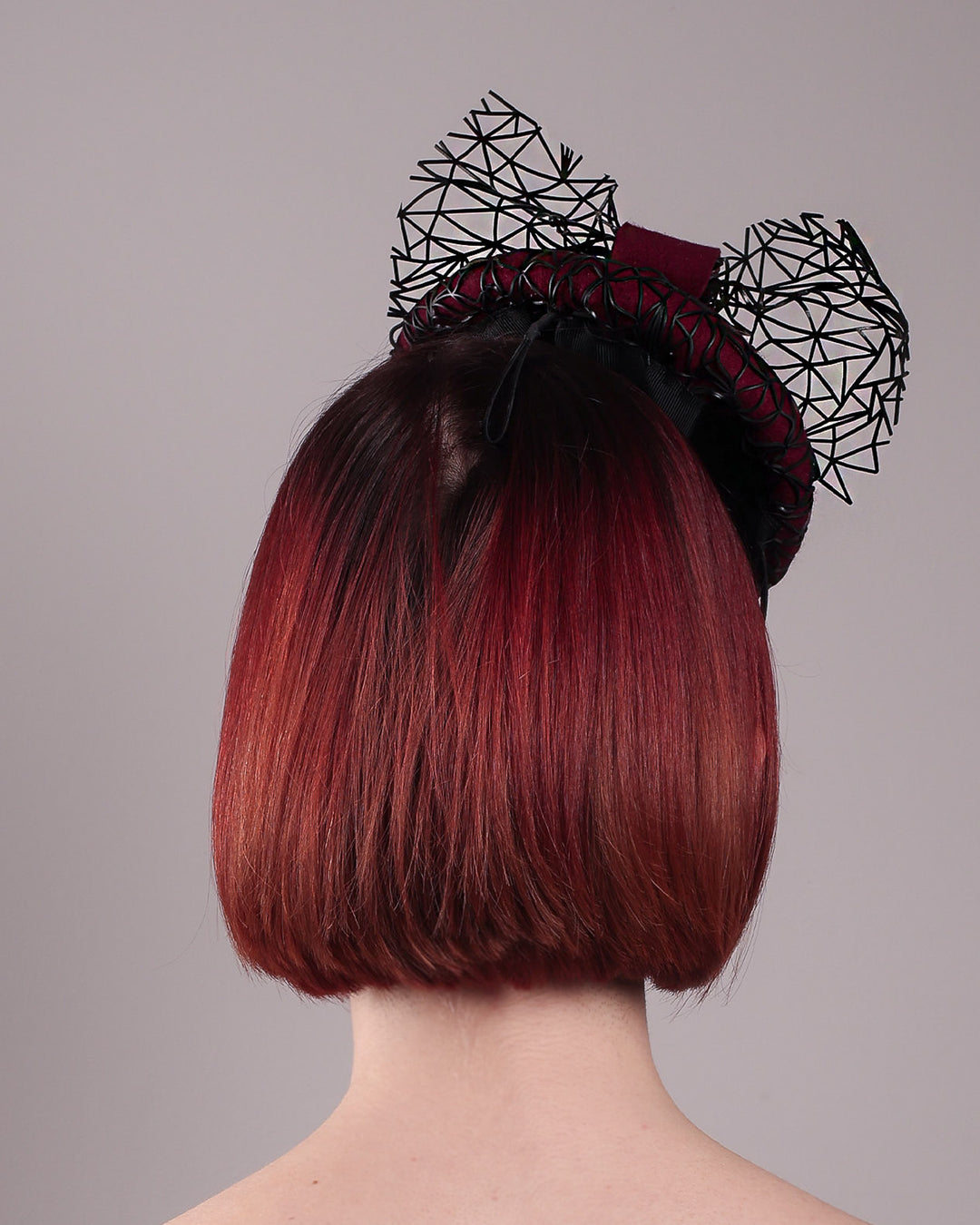 The model showcases the back of Bijou, featuring a handcrafted raffia fascinator in rich burgundy felt, with button shape. Its structured base is overlaid with an angular black netting design and crowned with an architectural bow that fuses avant-garde with elegance. Designed by Merve Bayindir.