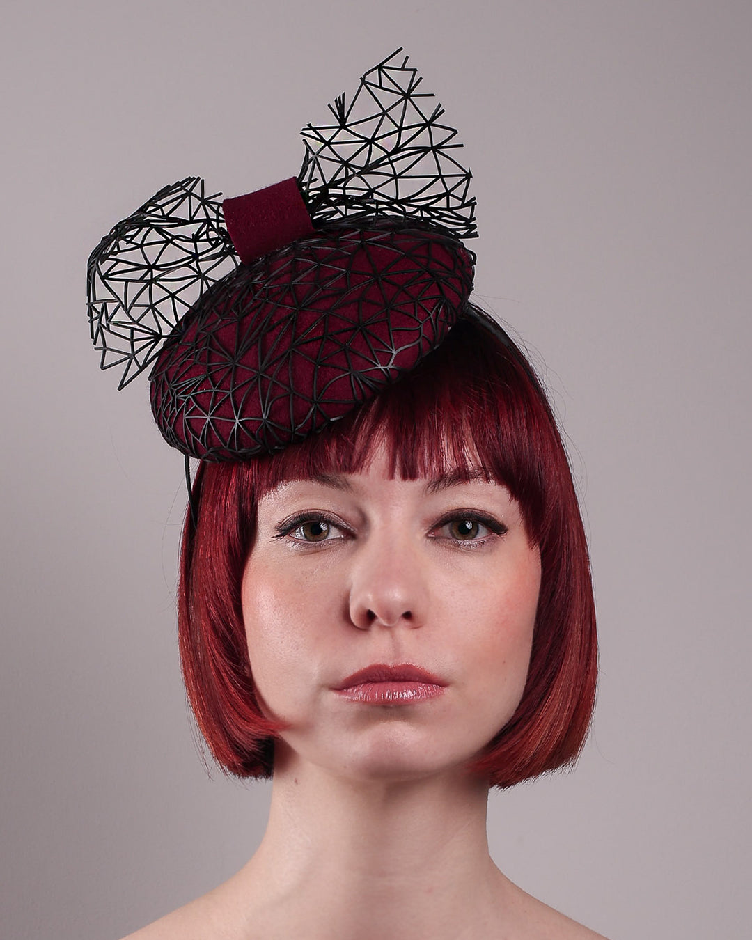 The model showcases the front close up of Bijou, featuring a handcrafted raffia fascinator in rich burgundy felt, with button shape. Its structured base is overlaid with an angular black netting design and crowned with an architectural bow that fuses avant-garde with elegance. Designed by Merve Bayindir.