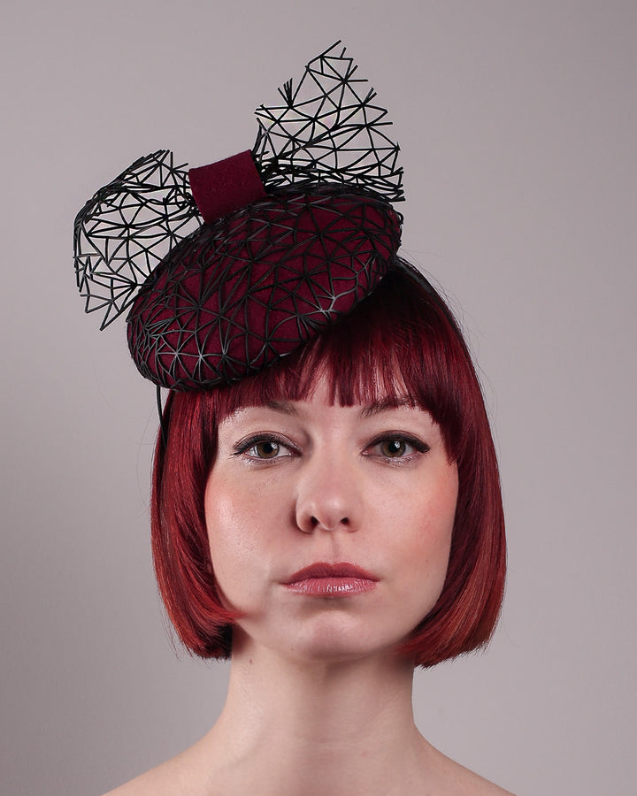 The model showcases the front close up of Bijou, featuring a handcrafted raffia fascinator in rich burgundy felt, with button shape. Its structured base is overlaid with an angular black netting design and crowned with an architectural bow that fuses avant-garde with elegance. Designed by Merve Bayindir.