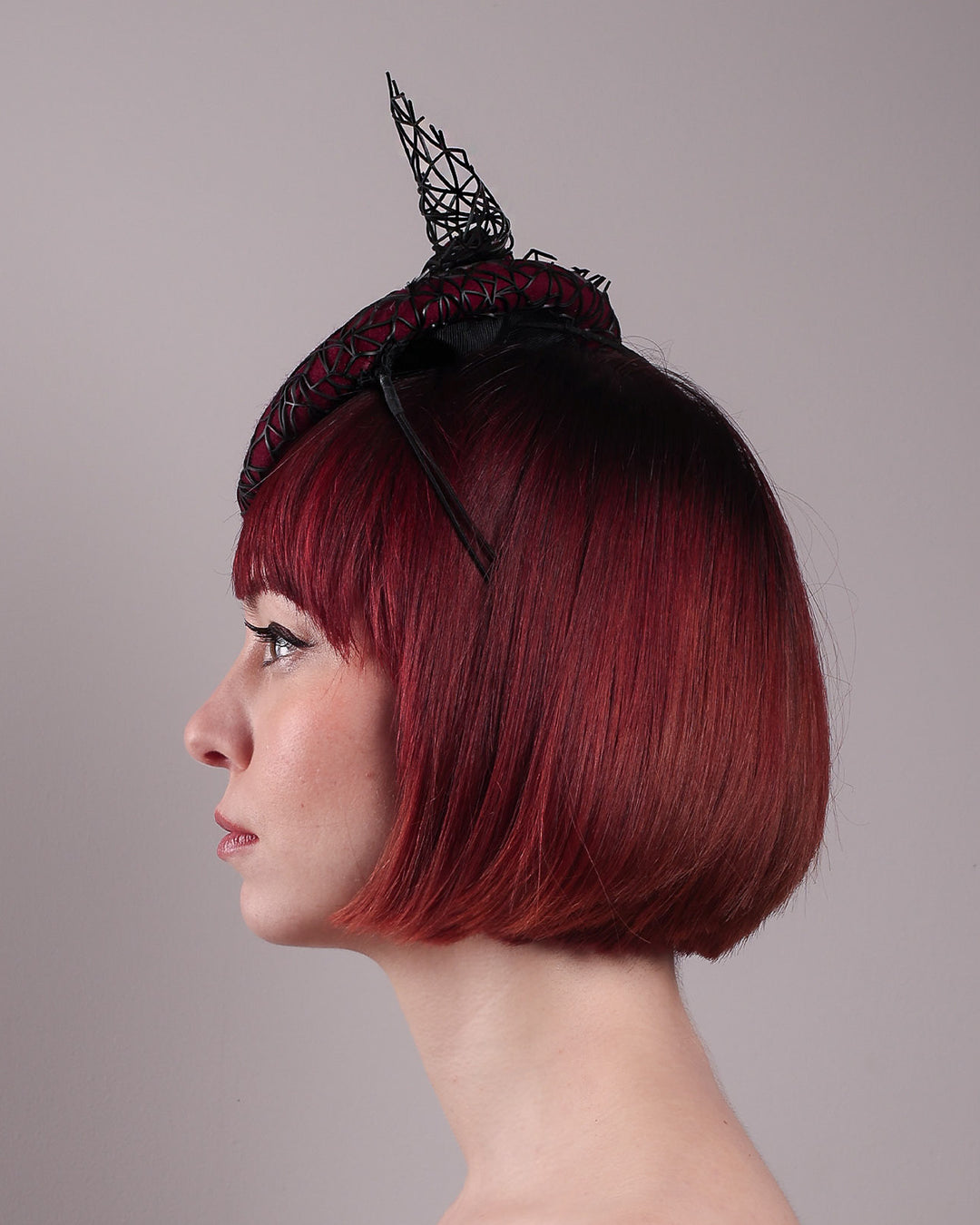 The model showcases the left side close up of Bijou, featuring a handcrafted raffia fascinator in rich burgundy felt, with button shape. Its structured base is overlaid with an angular black netting design and crowned with an architectural bow that fuses avant-garde with elegance. Designed by Merve Bayindir.