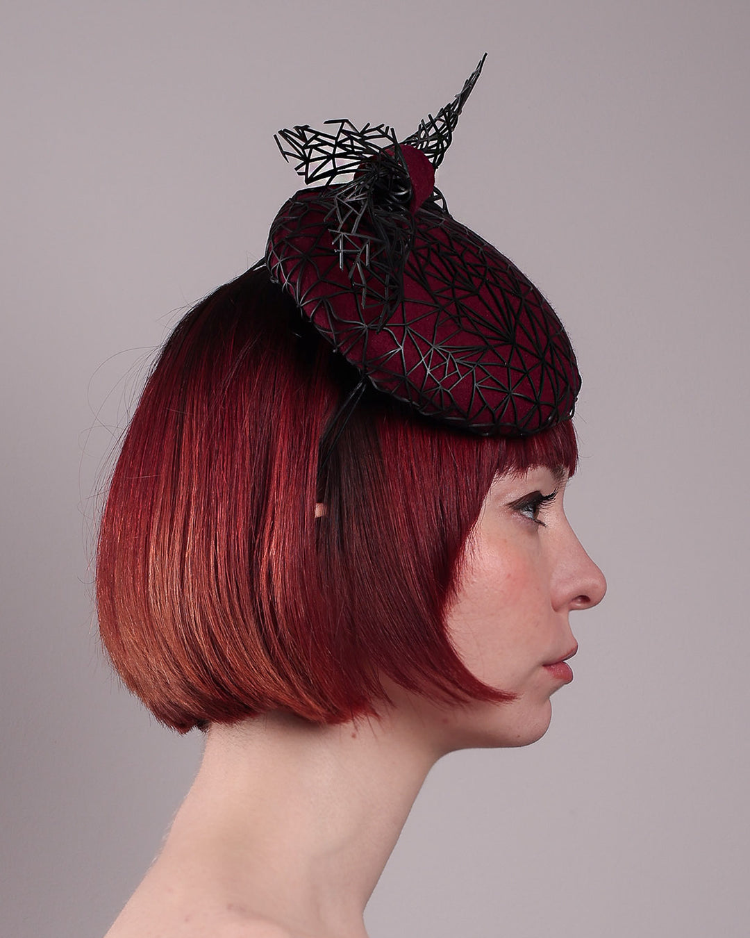 The model showcases the left side close up of Bijou, featuring a handcrafted raffia fascinator in rich burgundy felt, with button shape. Its structured base is overlaid with an angular black netting design and crowned with an architectural bow that fuses avant-garde with elegance. Designed by Merve Bayindir. 