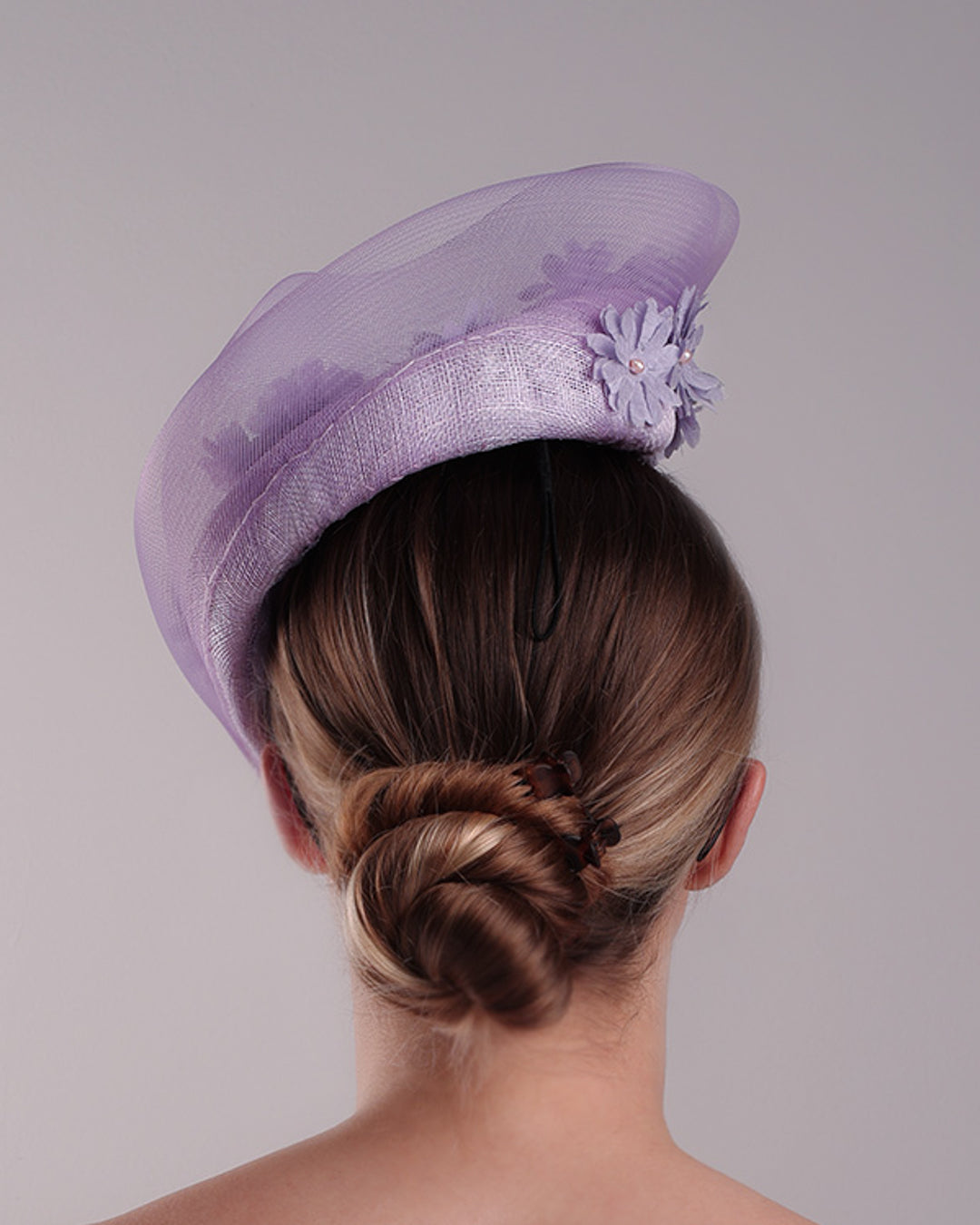 The model is showcasing the back of a lilac preacher style teardrop fascinator worn to the left side of the face. The piece drops from the top right of the head all the way the the right cheek like a teardrops. The hat from one side to the other from the back decorated with two layers of crinoline creating a wave effect. Ebellished with silk taffeta flowers.