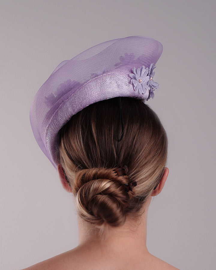 The model is showcasing the back of a lilac preacher style teardrop fascinator worn to the left side of the face. The piece drops from the top right of the head all the way the the right cheek like a teardrops. The hat from one side to the other from the back decorated with two layers of crinoline creating a wave effect. Ebellished with silk taffeta flowers.