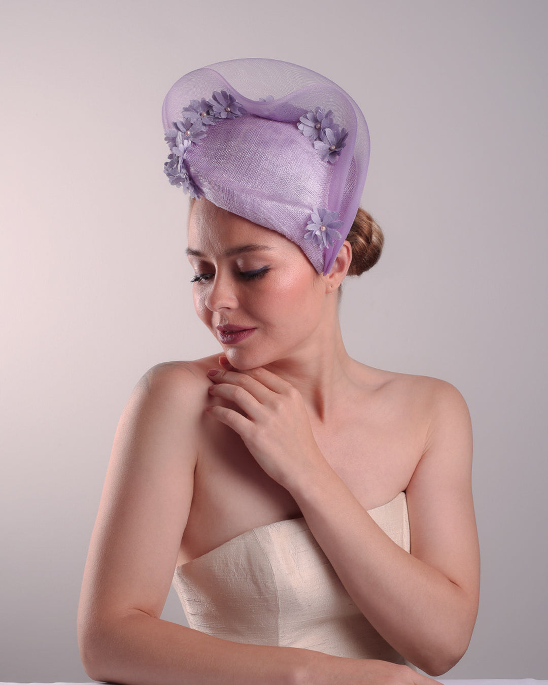 The model is showcasing the left front side of a lilac preacher style teardrop fascinator worn to the left side of the face. The piece drops from the top right of the head all the way the the right cheek like a teardrops. The hat from one side to the other from the back decorated with two layers of crinoline creating a wave effect. Ebellished with silk taffeta flowers.
