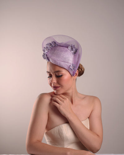 The model is showcasing the left front side of a lilac preacher style teardrop fascinator worn to the left side of the face. The piece drops from the top right of the head all the way the the right cheek like a teardrops. The hat from one side to the other from the back decorated with two layers of crinoline creating a wave effect. Ebellished with silk taffeta flowers.