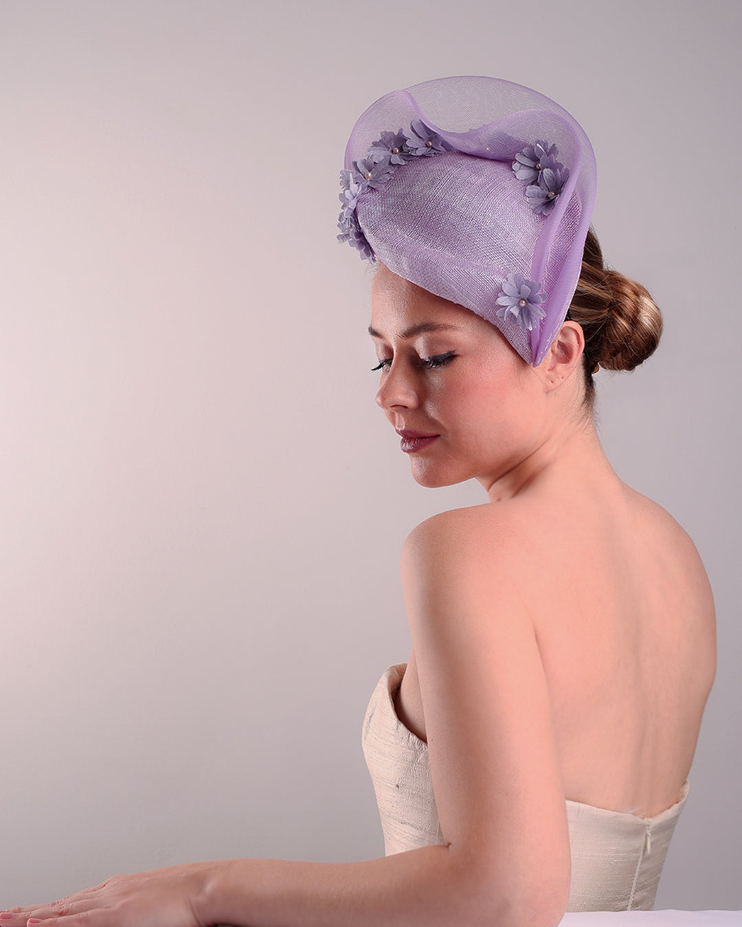 The model is showcasing the left front side of a lilac preacher style teardrop fascinator worn to the left side angle of the face. The piece drops from the top right of the head all the way the the right cheek like a teardrops. The hat from one side to the other from the back decorated with two layers of crinoline creating a wave effect. Ebellished with silk taffeta flowers.