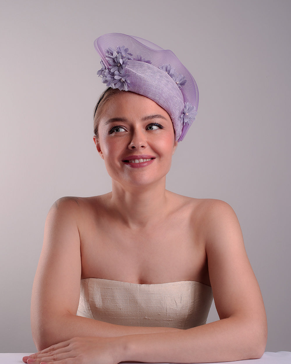 The model is showcasing the front of a lilac preacher style teardrop fascinator worn to the left side of the face. The piece drops from the top right of the head all the way the the right cheek like a teardrops. The hat from one side to the other from the back decorated with two layers of crinoline creating a wave effect. Ebellished with silk taffeta flowers.