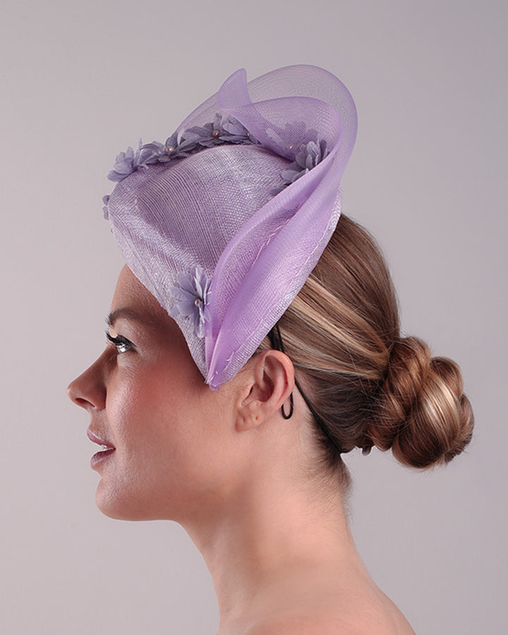 The model is showcasing the left side of a lilac preacher style teardrop fascinator worn to the left side of the face. The piece drops from the top right of the head all the way the the right cheek like a teardrops. The hat from one side to the other from the back decorated with two layers of crinoline creating a wave effect. Ebellished with silk taffeta flowers.