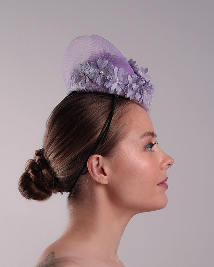 The model is showcasing the right side of a lilac preacher style teardrop fascinator worn to the left side of the face. The piece drops from the top right of the head all the way the the right cheek like a teardrops. The hat from one side to the other from the back decorated with two layers of crinoline creating a wave effect. Ebellished with silk taffeta flowers.
