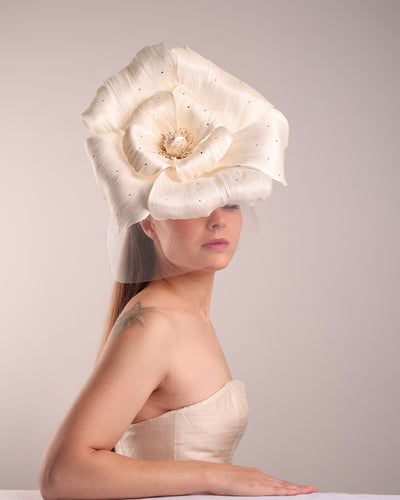 Model is showcasing a large white hand made silk abaca flower, decorated with silver crystals, placed one a button shaped white sinamay fascinator. A white crinoline veil is placed between the hat and the flower to cover the face. The button base is fixed on a custom headband and secured with an additional elastic.