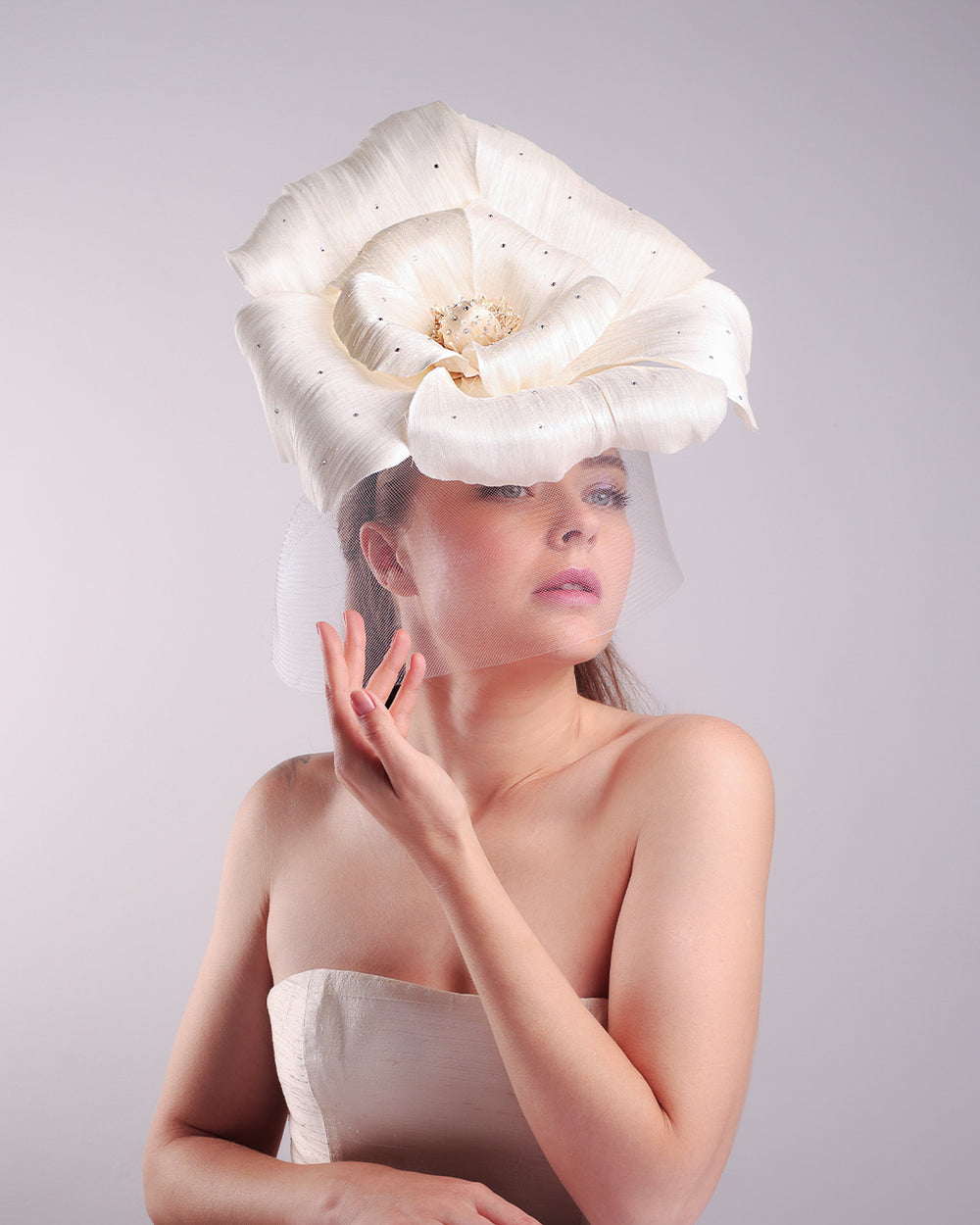Model is showcasing a large white hand made silk abaca flower, decorated with silver crystals, placed one a button shaped white sinamay fascinator from the right front side. A white crinoline veil is placed between the hat and the flower to cover the face. The button base is fixed on a custom headband and secured with an additional elastic.