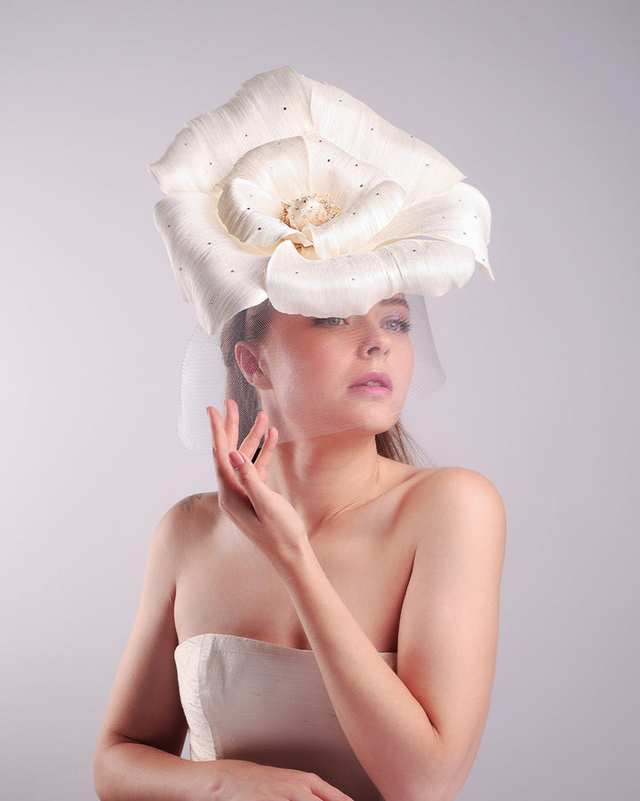 Model is showcasing a large white hand made silk abaca flower, decorated with silver crystals, placed one a button shaped white sinamay fascinator from the right front side. A white crinoline veil is placed between the hat and the flower to cover the face. The button base is fixed on a custom headband and secured with an additional elastic.