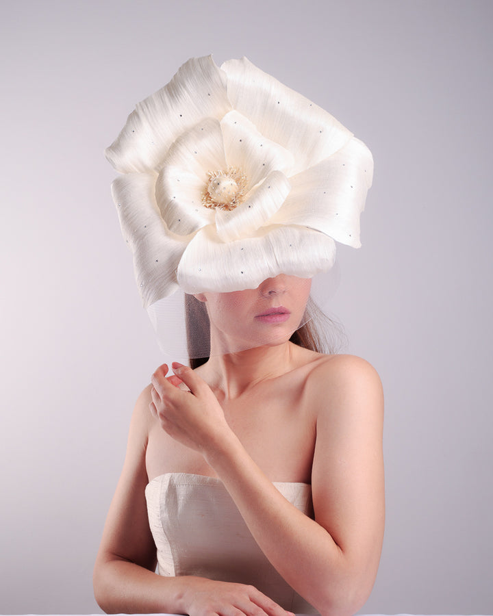 Model is showcasing a large white hand made silk abaca flower, decorated with silver crystals, placed one a button shaped white sinamay fascinator from the above focusing on the flowers. A white crinoline veil is placed between the hat and the flower to cover the face.