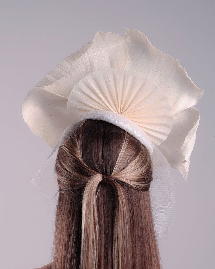 Model is showcasing the back of a large white hand made silk abaca flower, decorated with silver crystals, placed one a button shaped white sinamay fascinator from the left side A white crinoline veil is placed between the hat and the flower to cover the face. The button base is fixed on a custom headband and secured with an additional elastic.