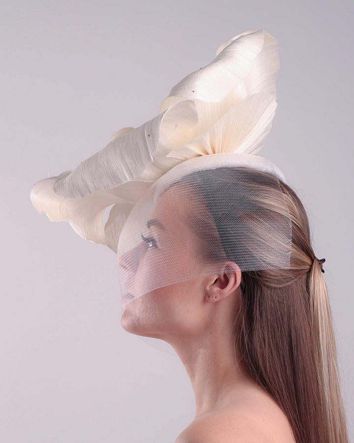 Model is showcasing a large white hand made silk abaca flower, decorated with silver crystals, placed one a button shaped white sinamay fascinator from the left side A white crinoline veil is placed between the hat and the flower to cover the face. The button base is fixed on a custom headband and secured with an additional elastic.