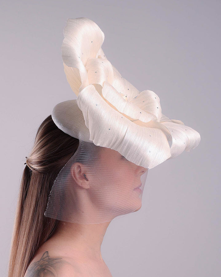 Model is showcasing the right side of a large white hand made silk abaca flower, decorated with silver crystals, placed one a button shaped white sinamay fascinator from the left side A white crinoline veil is placed between the hat and the flower to cover the face. The button base is fixed on a custom headband and secured with an additional elastic.