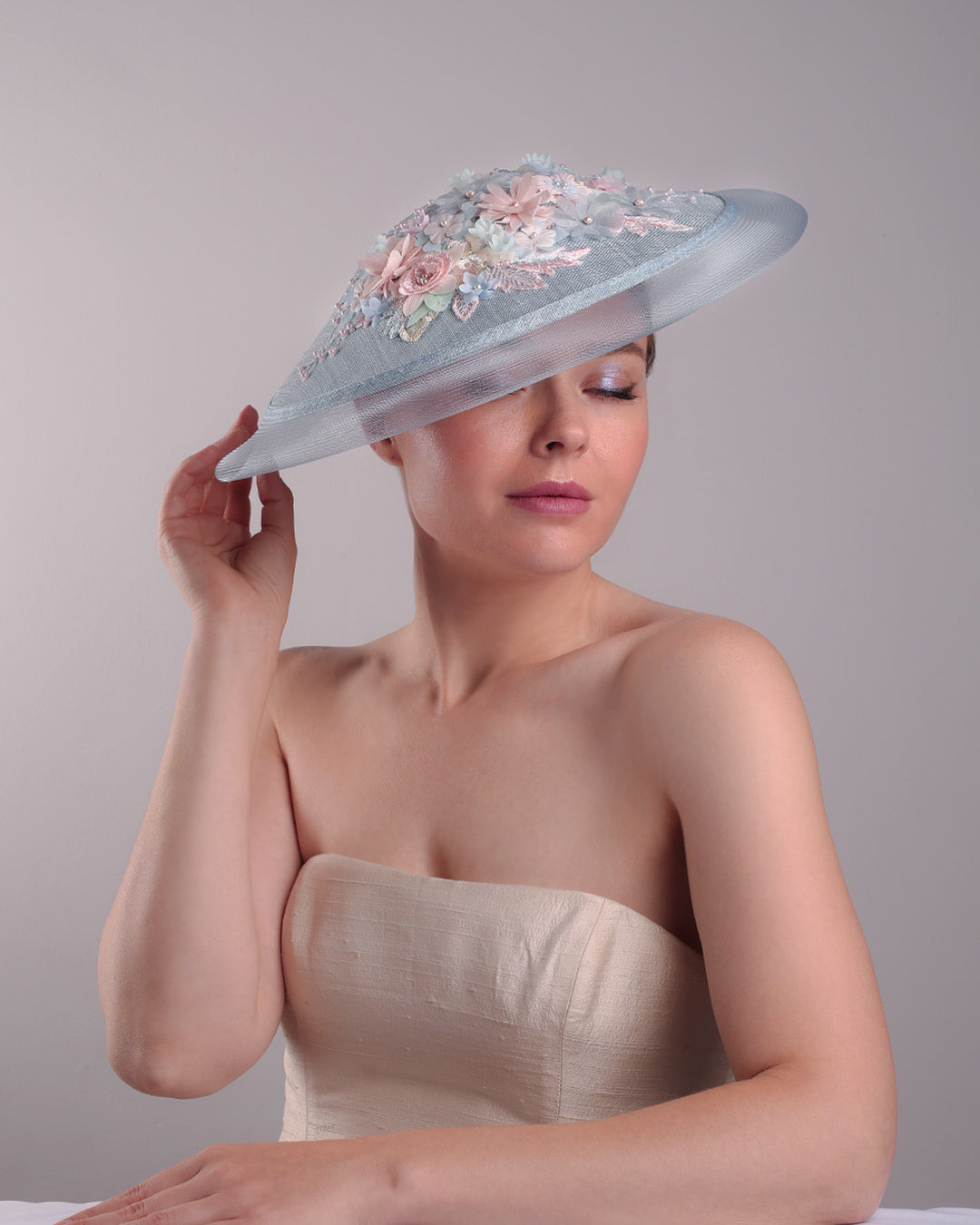 Model is showcasing the front right angle view of a blue circle style saucer hat, extended with crinoline, decorated on the top with a 3D embroidery that is embellised with pearls, and silk flowers. The hat is worn tilted to the right side. The hat is fitted to the head with a wire headband and elastic.