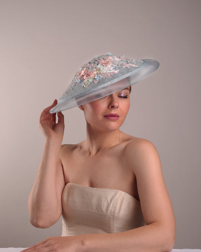 Model is showcasing the front right angle view of a blue circle style saucer hat, extended with crinoline, decorated on the top with a 3D embroidery that is embellised with pearls, and silk flowers. The hat is worn tilted to the right side. The hat is fitted to the head with a wire headband and elastic.