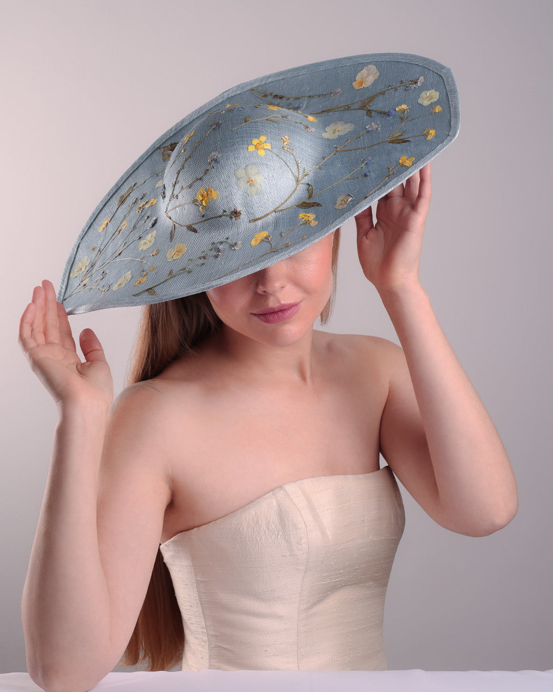 The model is showcasing the top angle of a blue asymetric whide brim hat, tilted to the right, decorated with pressed natural flowers creatinga almost 3D dimensional print affect on the hat. Fitted with a wired headband and additionally secured with elastic.
