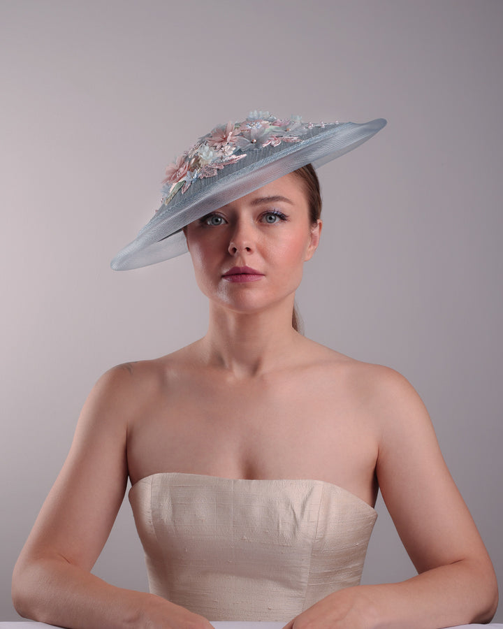 Model is showcasing the front view of a blue circle style saucer hat, extended with crinoline, decorated on the top with a 3D embroidery that is embellised with pearls, and silk flowers. The hat is worn tilted to the right side. The hat is fitted to the head with a wire headband and elastic.